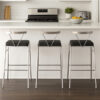 Three stainless steel kitchen bar chairs with black seats lined up at a white kitchen island